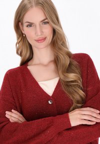 Czerwony kardigan z dekoltem w serek, długimi rękawami i srebrnym detalem z guzikiem. Miękka tekstura z subtelnymi akcentami blasku. Noszony na kremową bluzkę.