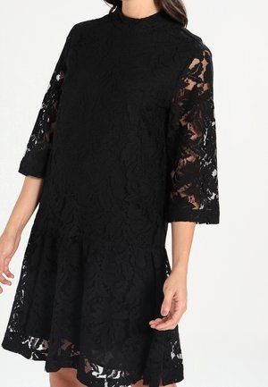 Femme portant une robe noire en dentelle avec des manches trois-quarts et un ourlet légèrement évasé, présentant des motifs floraux en dentelle et un col montant.