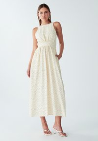 Vestido maxi en crema con lunares negros, que presenta un cuerpo ajustado, cintura fruncida y falda relajada. Fabricado con tejido ligero.