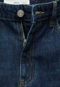 Close-up van donkerblauwe denim jeans met gedeeltelijk opengeritste zilveren rits, metalen knop, zichtbare riemlus en gestikte naden.