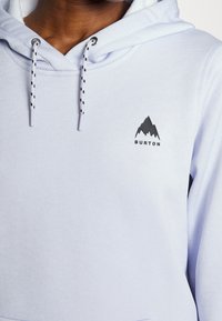 Personne portant un sweat à capuche violet clair avec des cordons noirs et blancs et un logo de montagne noir avec le texte "BURTON" sur la poitrine.