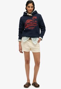 Haut à capuche bleu marine avec logo rouge, chemise rayée bleu clair en dessous, shorts blancs effilochés et sandales marron. Design simple et coupe décontractée.
