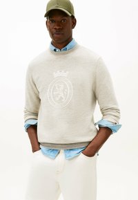Mand iført beige sweatshirt med løveemblem over lys blå skjorte, cremefarvede bukser og olivengrøn cap, hænderne i lommerne mod hvid baggrund.