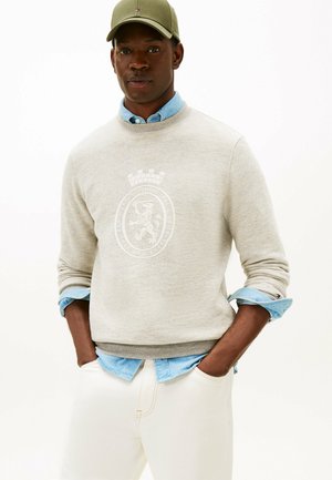 Man wearing beige sweatshirt with lion emblem over light blue shirt, cream pants, and olive green cap, hands in pockets against white background.