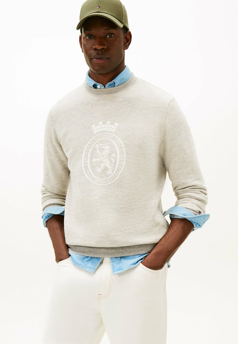 Mand iført beige sweatshirt med løveemblem over lys blå skjorte, cremefarvede bukser og olivengrøn cap, hænderne i lommerne mod hvid baggrund.