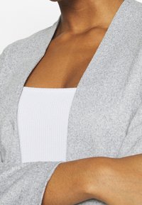 Harmaa neulecardigan, jossa on pehmeä, tekstuurinen pinta, avonaisella etuosalla ja leveillä hihoilla, päällekkäin valkoisen röyhelötoppi kanssa.