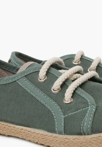 Scarpe in tela verde con lacci crema, occhielli in metallo e suole intrecciate in juta. Presentano una cucitura a contrasto lungo i bordi.