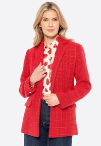 Blazer rosso testurizzato con una vestibilità su misura, caratterizzato da un motivo a quadri, rever a punta e tasche frontali. Indossato sopra una blusa floreale.