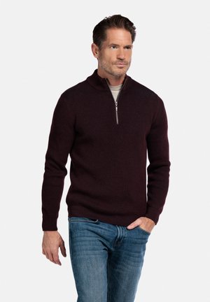 Pull en ribbed bordeaux avec un col montant et une fermeture éclair, associé à un jean bleu. Accent sur la texture et le design ajusté.