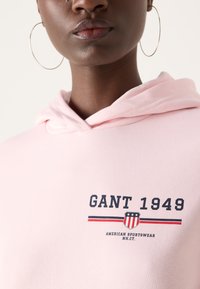 Ružová mikina s kapucňou na šnúrku, s logom "GANT 1949" v námorníckej a červenej pruhovanej kombinácii, vyrobená z mäkkého bavlneného materiálu.