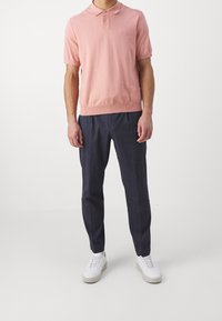 Uomo con una polo rosa a maniche corte, pantaloni blu scuro e sneakers bianche in piedi contro uno sfondo neutro.