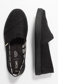 Svarta slip-on skor med ett strukturerat textilöverdrag, elastiska sidodetaljer och en solid svart gummisula. Inuti har de ett randigt mönster.