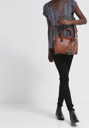 Femme portant un haut à manches courtes à motifs, un pantalon noir et des bottes, portant un sac à main en cuir marron avec des franges sur l'épaule.