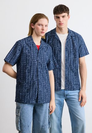 Giovane donna e uomo in piedi fianco a fianco, entrambi con magliette blu navy abbinate con scritte bianche in corsivo; la donna indossa jeans cargo, l'uomo jeans azzurri.