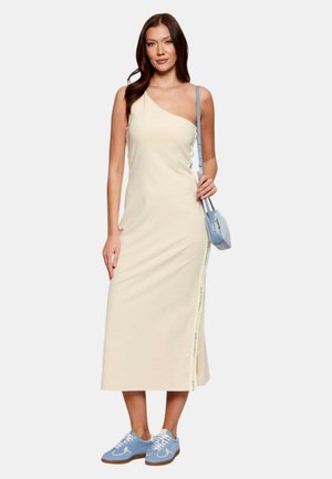 Donna che indossa un vestito midi crema monospalla, sneakers azzurro chiaro e una borsa a tracolla abbinata azzurro chiaro, in piedi davanti a uno sfondo semplice.