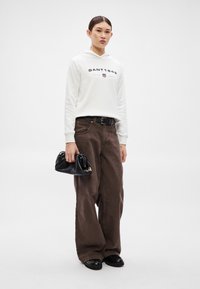 Hanorac alb cu inscripția "GANT 1949" pe partea din față, asortat cu pantaloni maro cu croială largă și pantofi negri comozi. Ține o geantă neagră.