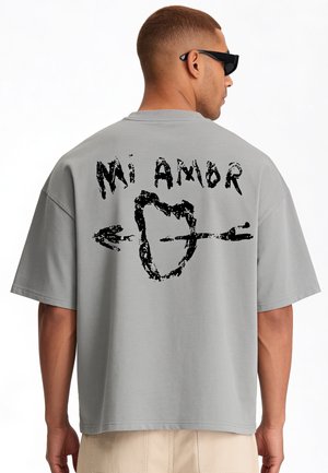 Man draagt een grijs oversized T-shirt met zwarte afbeelding en tekst "Mi Amor" op de rug, gecombineerd met beige broek en zwarte zonnebril.