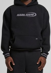 Černá mikina s kapucí s poněkud stříbrným logem "KARL KANI", žebrovanými manžetami a lemem s decentním pruhovaným detailem a přední klokaní kapsou.