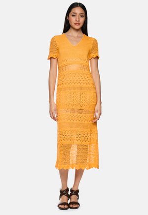 Woman wearing a mustard yellow crochet midi dress with short sleeves and black strappy sandals, standing against a plain background.