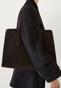Brązowa skórzana torba typu tote o minimalistycznym designie, prostokątnym kształcie i długich uchwytach, wyeksponowana obok czarnego płaszcza z szerokimi rękawami.