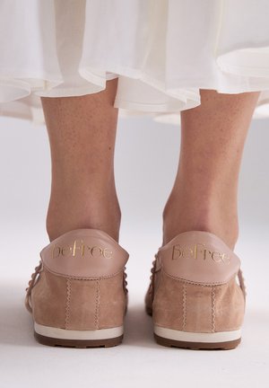 Vista trasera de los pies con zapatillas de gamuza beige y texto dorado "BeFree" en las lengüetas del talón, bajo una falda blanca fluida.