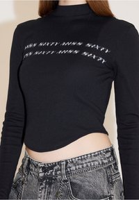 Top negro recortado de manga larga con cuello redondo, que presenta texto reflectante en el pecho, combinado con jeans de mezclilla desgastados de tiro alto.
