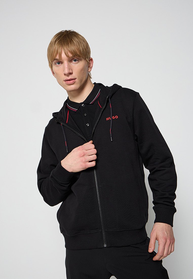 Schwarze Zip-Hoodie mit Kapuze, roten Akzentnähten und "HUGO"-Logo auf der Brust. Das Model trägt ein schwarzes Polo darunter.