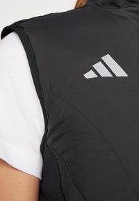 Černá bezrukávová vesta z hladkého materiálu, s šedým logem Adidas na levém rameni a kontrastním bílým tričkem pod ní.