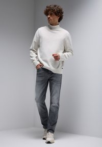 Lichtgrijze coltrui van zacht materiaal, gecombineerd met donkergrijze jeans. Model draagt neutrale sneakers en staat in een minimalistische omgeving.