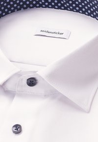 Witte overhemden met een klassieke kraag en donkerblauwe knopen, gemarkeerd met "seidensticker," op een donkerblauwe stof met lichtblauwe stippen.