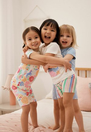 Three young children smiling and hugging each other while standing on a bed in a softly lit bedroom.