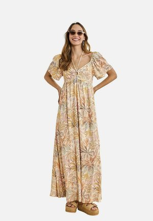 Mujer con un vestido largo de estampado floral, mangas abullonadas, detalle de anillo en el busto, gafas de sol, pendientes y sandalias de plataforma, sonriendo.