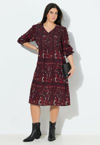 Burlrot Kleid mit V-Ausschnitt und langen Puffärmeln, das ein wirbelndes abstraktes Muster aufweist. Kombiniert mit schwarzen Absatzstiefeln und einer Clutch.