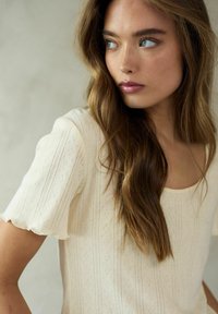 Krämfärgad topp med korta, scallopformade ärmar. Har vertikala ränder och ett texturerat mönster. Rundad halsringning och figurnära silhuett.