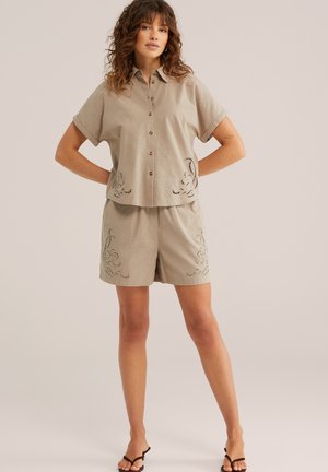 Donna in piedi con le mani sui fianchi, indossa una camicia beige a maniche corte con bottoni e pantaloncini abbinati con disegni ricamati ritagliati.