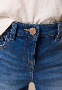 Modre denim hlače imajo visok pas, z mesinganimi gumbi in kontrastnim oranžnim šivanjem, s prednjimi žepi, ki so vidni.