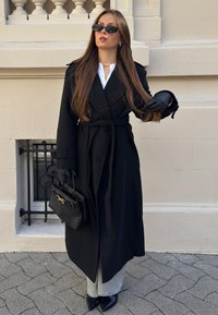 Svart trenchcoat med bälte, bärs över en vit skjorta. Svarta läderhandskar, svart vävd handväska och spetsiga svarta stövlar fulländar looken.