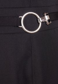 Boucle circulaire en métal argenté avec gravure "HUGO" attachée à une ceinture en tissu noir sur des vêtements noirs texturés.