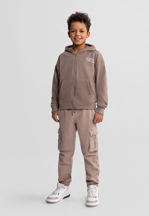 Młody chłopiec stojący, ubrany w szarobrązową bluzę na zamek, dopasowane spodnie typu cargo i białe wysokie trampki, uśmiechający się do kamery.