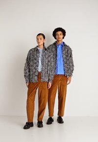 Dois modelos a usar jaquetas cinza texturizadas correspondentes, calças castanhas e sapatos pretos, em pé contra um fundo branco liso.