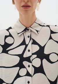 Valge särk abstraktsete mustade kujunditega, nööpidega krae ja mustade nööpidega. Omab tekstuurset kangast, millel on kortsus välimus.