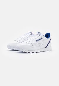 Pair of white Reebok Classic leather sneakers with blue interior lining and logo on side and tongue, set against white background.