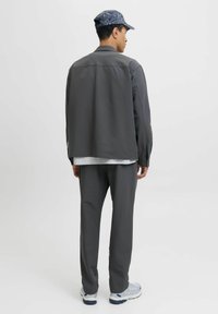Jonge man in een grijze jas, bijpassende broek, witte sneakers en een blauw patroonpet, staand met zijn rug naar de camera op een effen lichte achtergrond.
