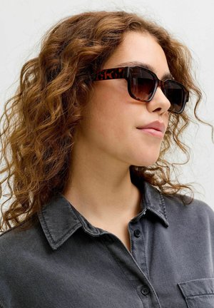 Kobieta o kręconych brązowych włosach, nosząca okulary przeciwsłoneczne w oprawkach w kolorze żółwia oraz ciemnoszarą koszulę z kołnierzykiem, zwrócona lekko w prawo na jasnym tle.