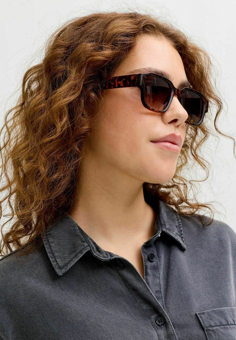 Frau mit lockigem braunem Haar, die Schildpatt-Sonnenbrillen und ein dunkelgraues Hemd mit Kragen trägt, leicht nach rechts gewandt vor hellem Hintergrund.