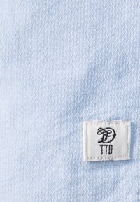 Ljust blått strukturerat tyg med ett litet vitt märke med svarta bokstäver "TTD". Materialet verkar slätt med ett subtilt mönster.