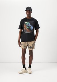 Schwarzes T-Shirt mit einem grafischen Druck eines Eidechsenmotivs, kombiniert mit beigen Shorts, die ein natur inspiriertes Muster zeigen, und hellbraunen Sneakers.