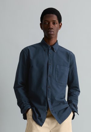 Junger Mann mit kurzen schwarzen Haaren, der ein dunkelblaues Hemd mit Knöpfen und beige Hosen trägt, steht mit den Händen in den Taschen vor einem grauen Hintergrund.