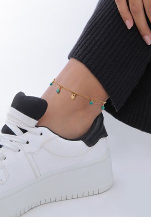 Goldene Fußkette mit Türkisperlen und einem Herzanhänger, getragen über einer schwarzen gerippten Socke und einem weißen Sneaker.