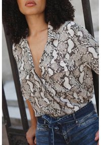 Vrouw in een shirt met slangenprint en hoog-taille blauwe jeans met een gestrikte riem, staand bij een zwart metalen hek.